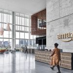 Corporate Amazon employee in office attire packing grocery orders inside a fulfillment warehouse during Prime Day peak, surrounded by automation equipment.