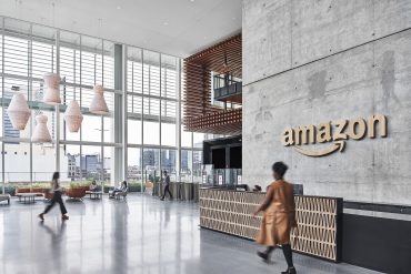 Corporate Amazon employee in office attire packing grocery orders inside a fulfillment warehouse during Prime Day peak, surrounded by automation equipment.