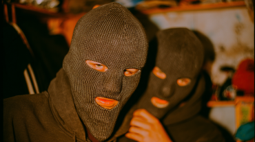 Two individuals wearing knitted gray balaclavas, standing close together in a dimly lit room.